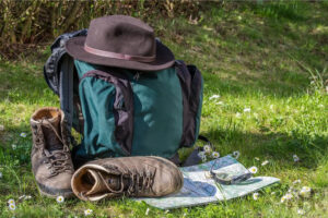 A green backpack, hiking boots, a brown hat, a map and a compass on a grassy field with small white flowers